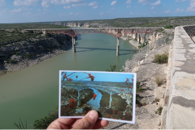 Hand holding old postcard of a bridge in front of the same bridge over a river canyon