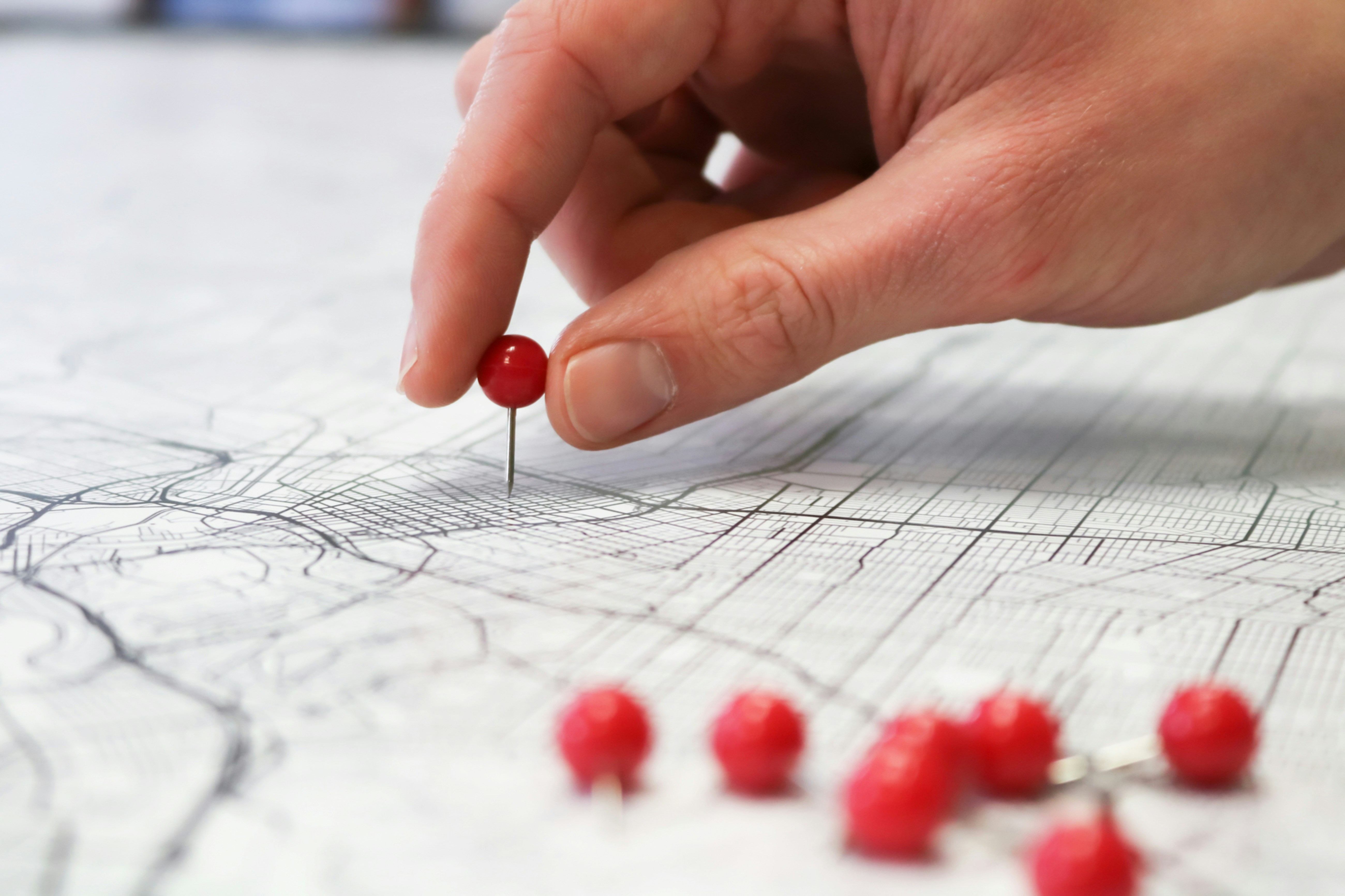 Hand placing a red pin on a detailed paper map with other pins nearby.